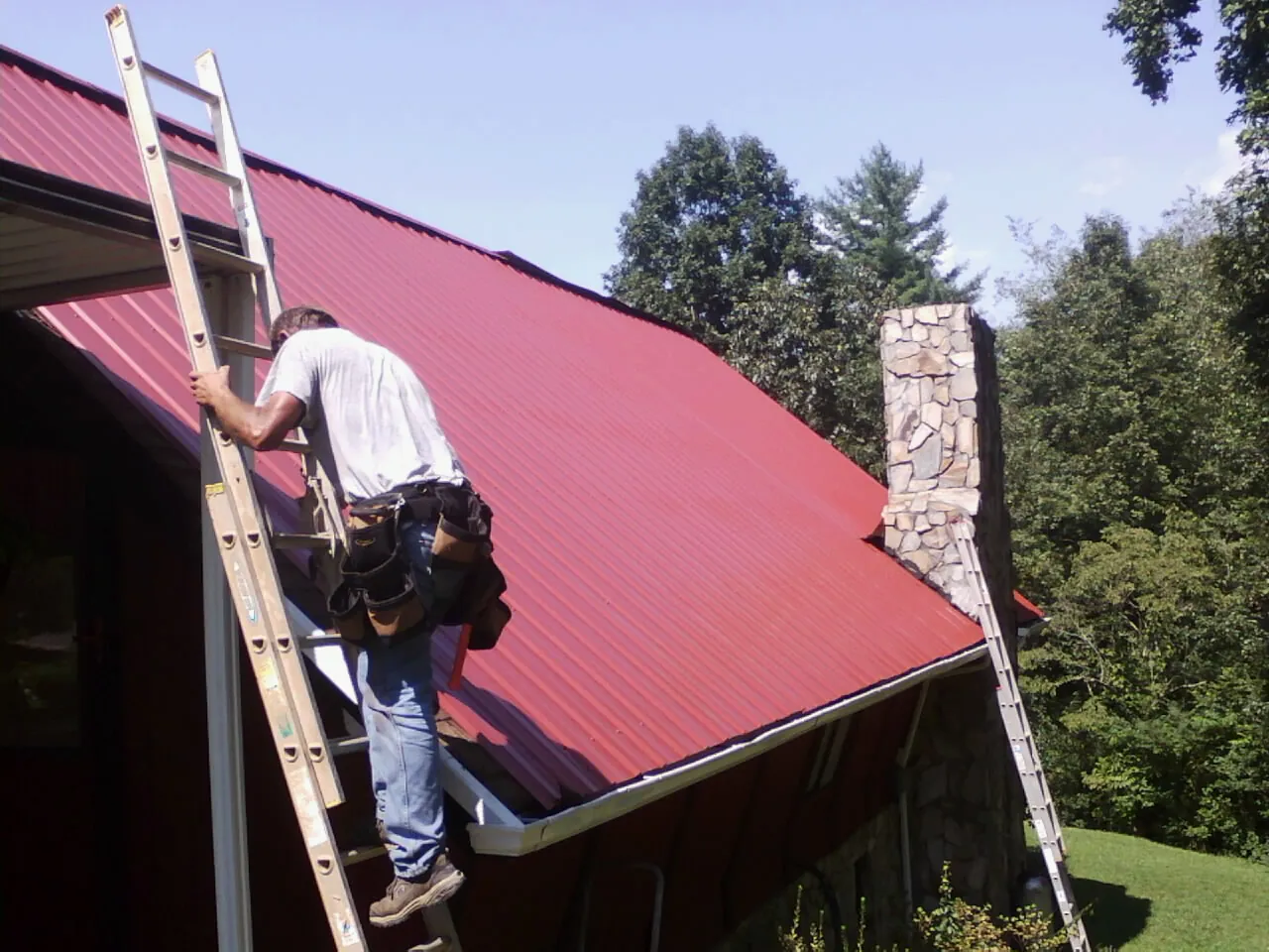 Roofer on a ladder beside a red metal roof and stone chimney — Soffit & Fascia Repair by Roofing Brothers in Molt